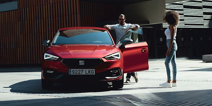 Vista frontal de hombre y mujer junto a un SEAT León en rojo desire en entorno urbano