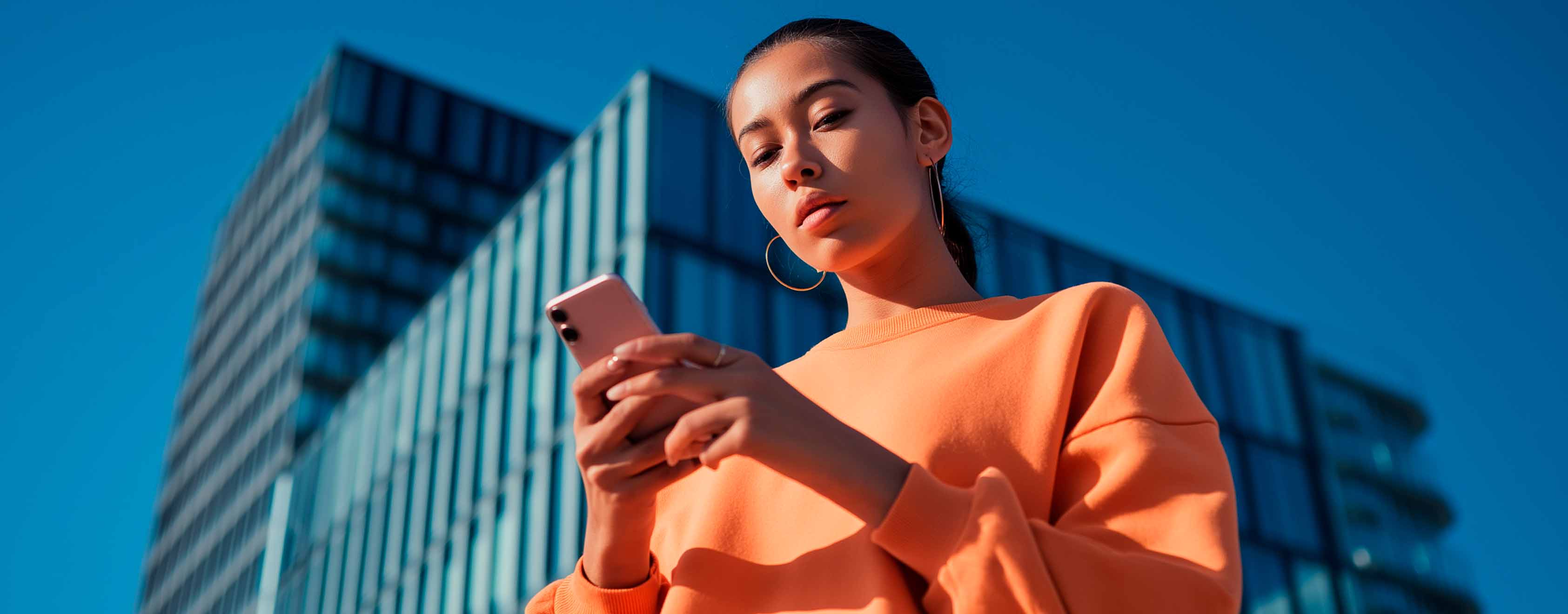 Mujer consulta smartphone; vista contrapicada frente a edificio de oficinas