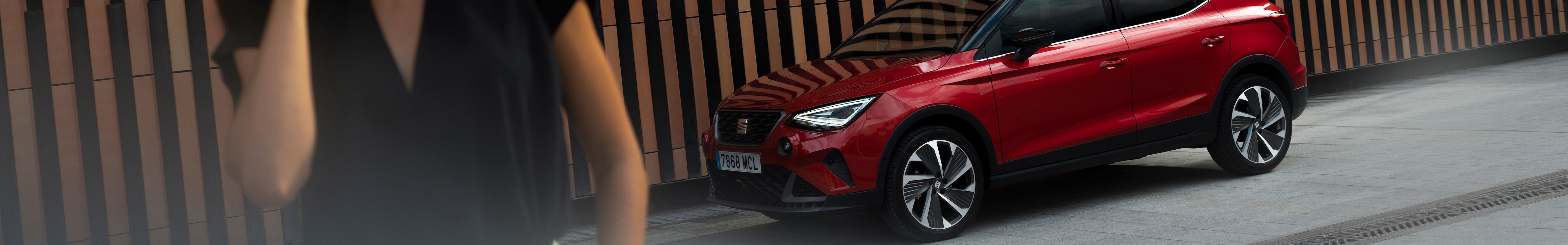 Mujer sonriendo camina con un SEAT Arona en color rojo desire al fondo