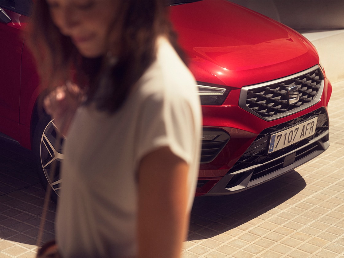 Parte delantera de un SEAT Ateca rojo aparcado en un lugar soleado. Una mujer está de pie junto al coche.