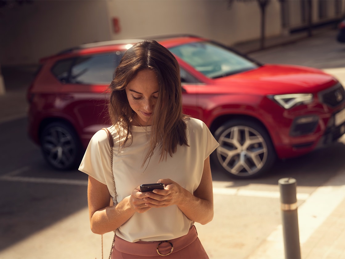 Mujer con smartphone utilizando los servicios online de SEAT Connect junto a un SEAT Ateca rojo.