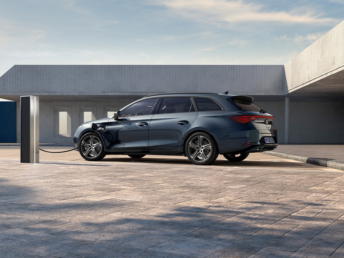 SEAT León Sportstourer compacto 2025, vehículo eléctrico híbrido enchufable y un cargador de SEAT. Estaciona y carga al aire libre, con llantas de aleación con radios, iluminación ambiental interior y asientos delanteros calefactados.