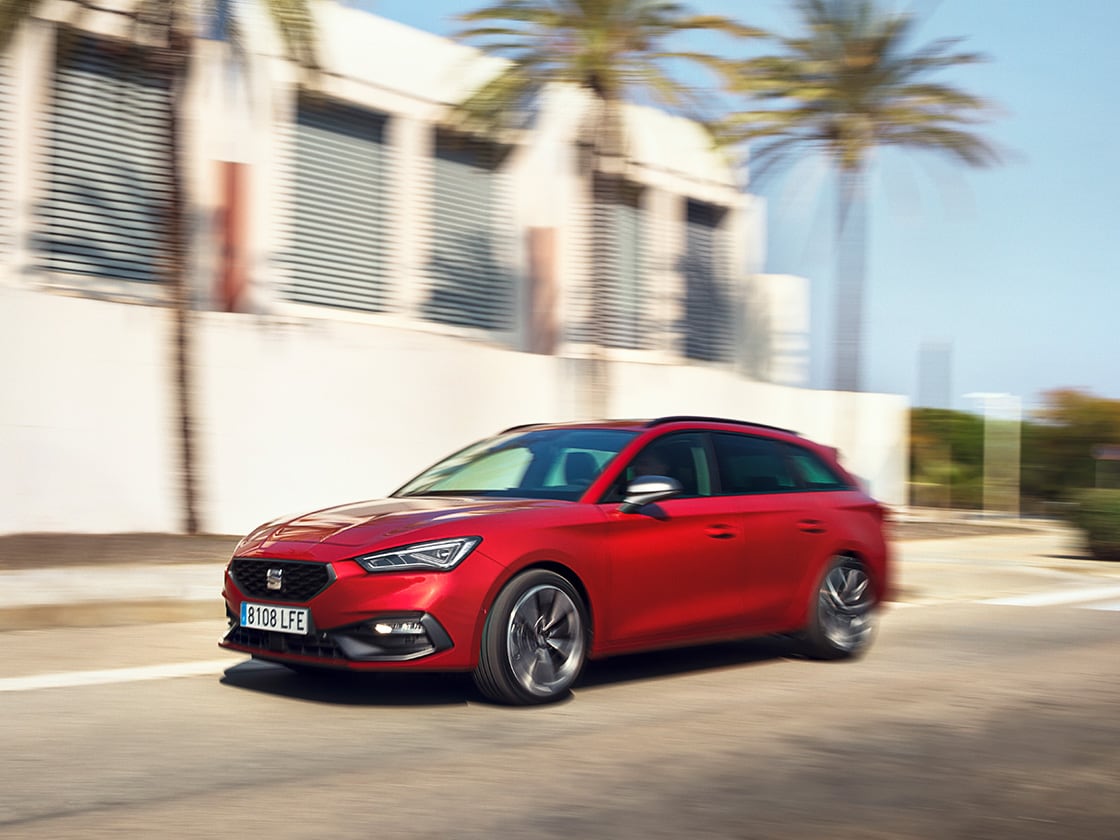 Un SEAT León Sportstourer rojo que disfruta del plan de servicio y mantenimiento de SEAT, en movimiento, en una zona residencial con una casa y palmeras.