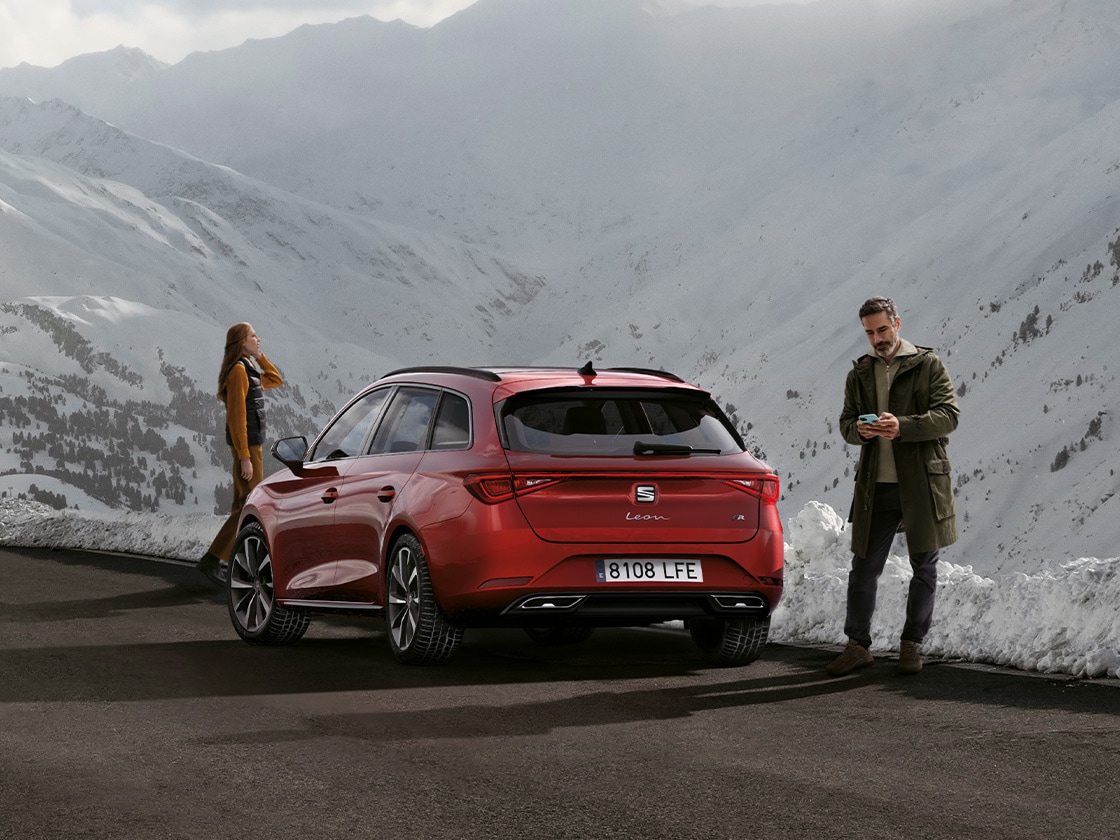 Un coche SEAT León Sportstourer rojo aparcado en una carretera de montaña esperando el servicio de SEAT Service Movilidad y asistencia en carretera. Dos personas de pie cerca.