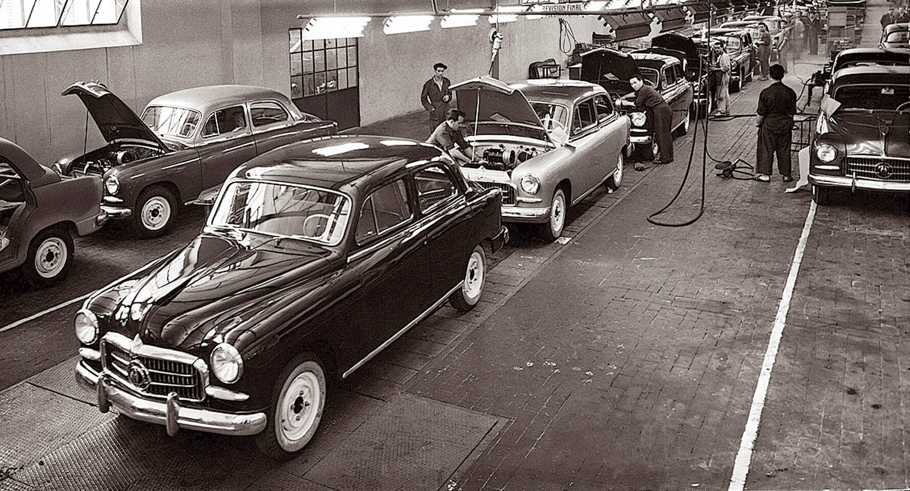 Línea de montaje de coches vintage con trabajadores y vehículos clásicos en una fábrica
