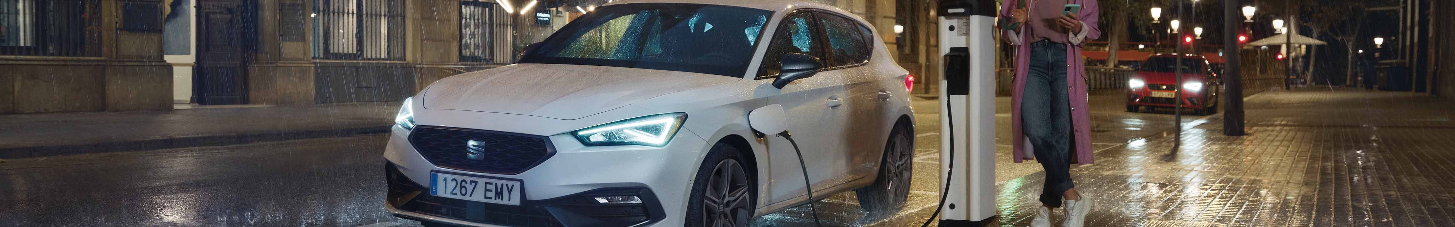 seat leon coche híbrido conectado a una estación de carga pública en una calle lluviosa de noche, resaltando las ventajas de los coches híbridos